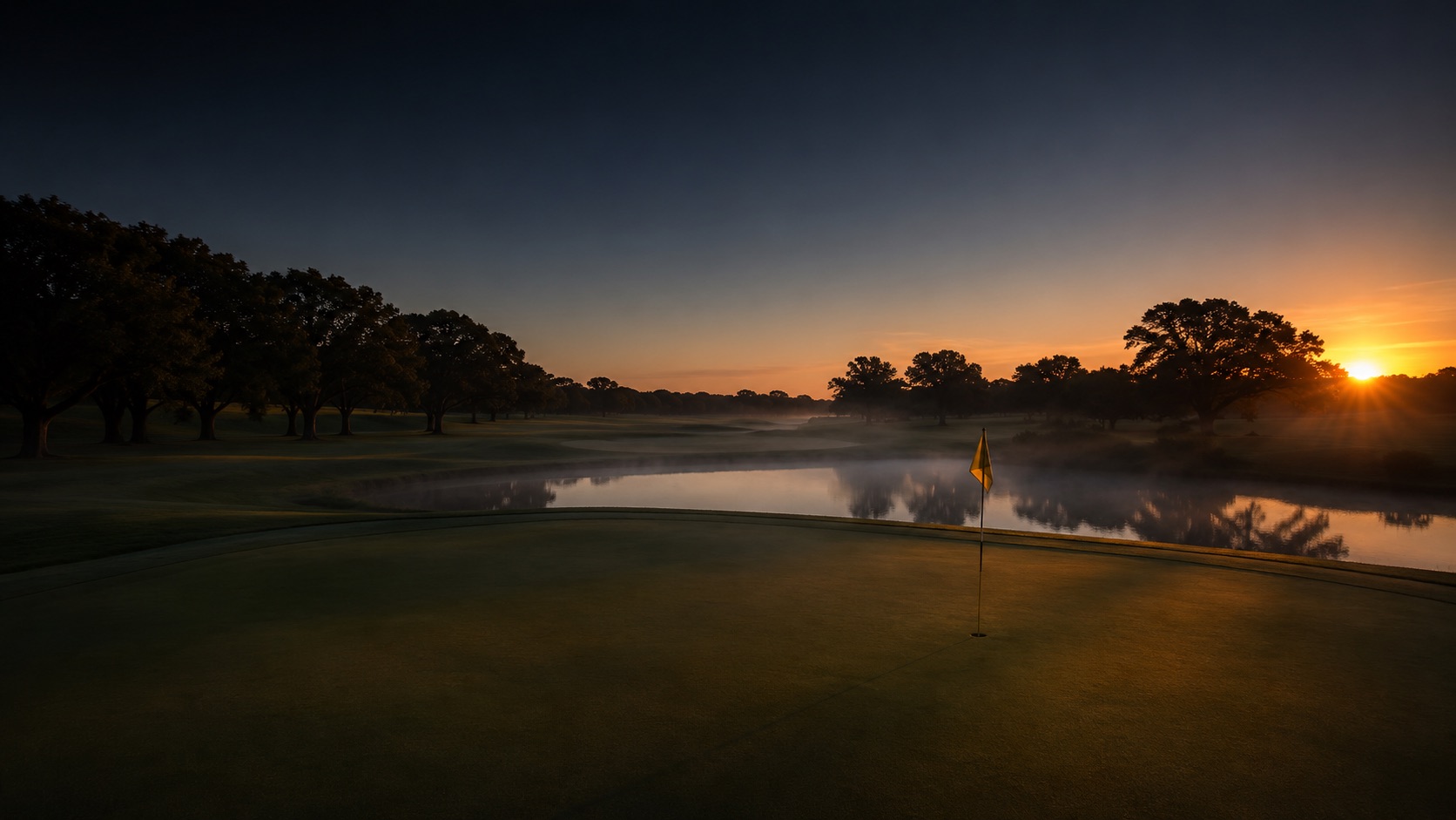 Sunrise over a championship golf course green with a yellow flag, calm water, and oak trees